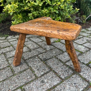Puede incluir: Un taburete de madera r&uacute;stico con una parte superior triangular y tres patas. El taburete est&aacute; hecho de madera toscamente labrada y tiene un acabado natural.