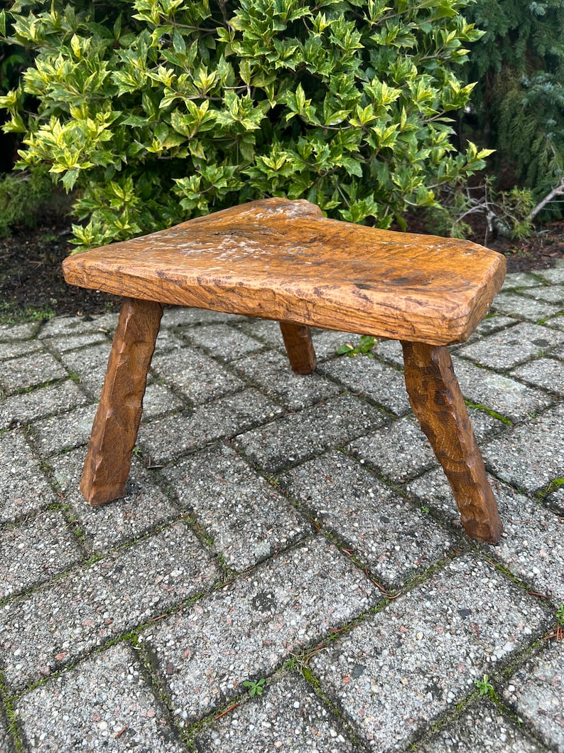 Puede incluir: Un taburete de madera r&uacute;stico con una parte superior triangular y tres patas. El taburete est&aacute; hecho de madera toscamente labrada y tiene un acabado natural.