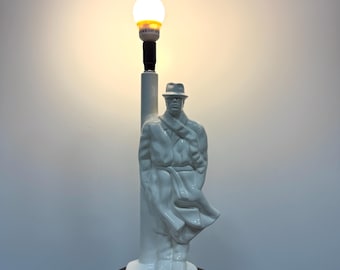 May include: White ceramic lamp with a man in a coat and hat standing on a post. The lamp is on a dark wood base.