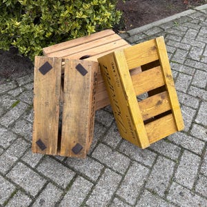 May include: Three wooden crates, one natural and two painted yellow, are arranged outdoors. The crates have a slatted design and metal corner reinforcements. One crate has the word "Holland" printed on it.