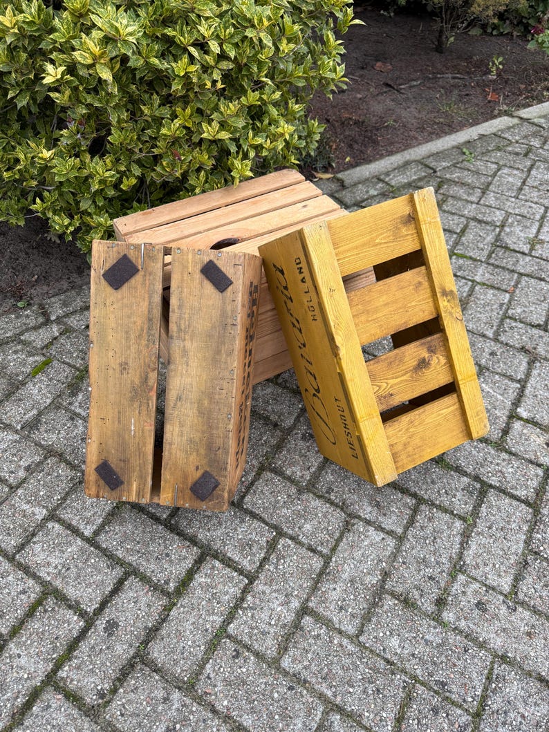 May include: Three wooden crates, one natural and two painted yellow, are arranged outdoors. The crates have a slatted design and metal corner reinforcements. One crate has the word "Holland" printed on it.