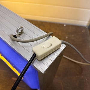 May include: Close-up of a rectangular, silver-colored metal object with a blue and yellow interior. A white on/off switch is attached to a black cord. A gray cord and a metal ring are also visible.