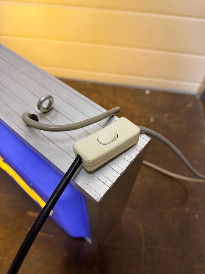 May include: Close-up of a rectangular, silver-colored metal object with a blue and yellow interior. A white on/off switch is attached to a black cord. A gray cord and a metal ring are also visible.