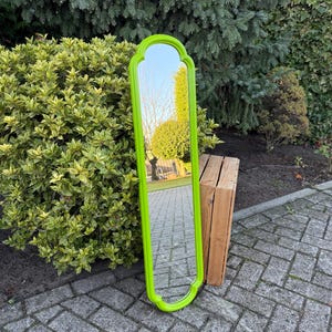 May include: A tall, rectangular mirror with a bright green frame. The mirror has a decorative, arched top and a curved bottom. The frame reflects the surrounding outdoor environment, including trees and sky.