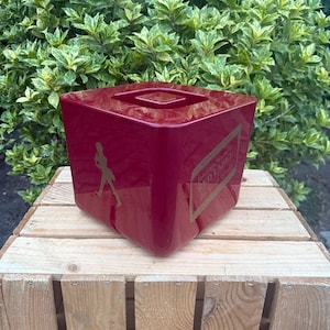 May include: A glossy, square, red container with a recessed top. The container features a gold label with the text "Johnnie Walker Red Label" and a silhouette of a striding man. The container is sitting on a wooden crate.