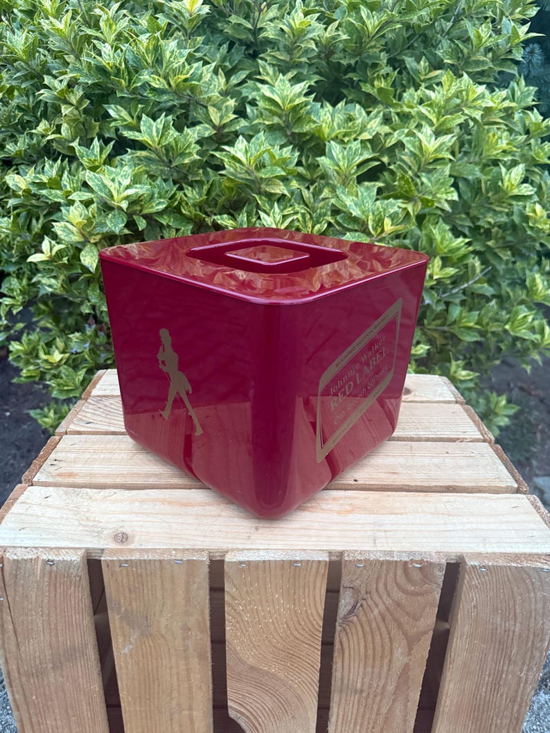 May include: A glossy, square, red container with a recessed top. The container features a gold label with the text "Johnnie Walker Red Label" and a silhouette of a striding man. The container is sitting on a wooden crate.