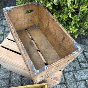 May include: A vintage wooden crate with metal corner brackets. The crate is rectangular with a handle cut-out. The wood has a natural, weathered look. The crate is placed on top of another wooden crate, with a third crate visible.