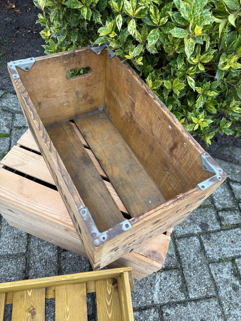 May include: A vintage wooden crate with metal corner brackets. The crate is rectangular with a handle cut-out. The wood has a natural, weathered look. The crate is placed on top of another wooden crate, with a third crate visible.