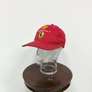 May include: A red Formula 1 baseball cap with a curved brim. The cap features the Formula 1 logo and a Ferrari emblem. The hat is displayed on a clear glass head form.