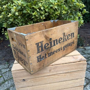 May include: A vintage wooden crate with the Heineken logo and text, placed on top of a smaller wooden crate. The crates are constructed from light brown wood and feature metal corner protectors. The text is in black.