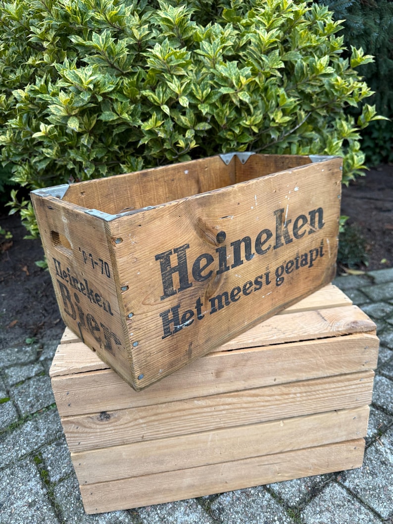May include: A vintage wooden crate with the Heineken logo and text, placed on top of a smaller wooden crate. The crates are constructed from light brown wood and feature metal corner protectors. The text is in black.