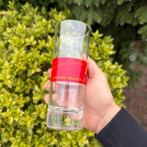 May include: A clear, tall glass with a red band featuring the text "JOHNNIE WALKER." The glass is held in a hand against a backdrop of green and yellow foliage. The glass is empty.