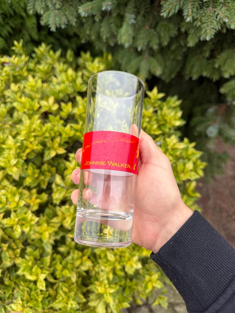 May include: A clear, tall glass with a red band featuring the text "JOHNNIE WALKER." The glass is held in a hand against a backdrop of green and yellow foliage. The glass is empty.