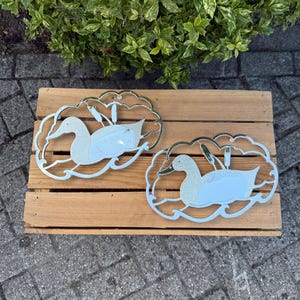 May include: Two decorative duck-shaped metal wall art pieces. Each duck is silver-coloured with a light blue body and a glittery silver head. The ducks are surrounded by a cloud-like frame with reeds.