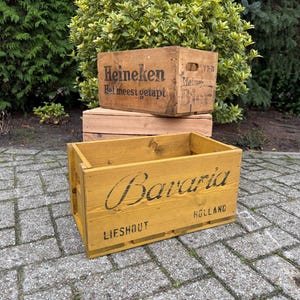 May include: Wooden crates with printed text. The top crate is light brown with the word "Heineken" and other text. The bottom crate is yellow with the word "Bavaria" and "Holland" printed on it. The crates are stacked on a brick surface.