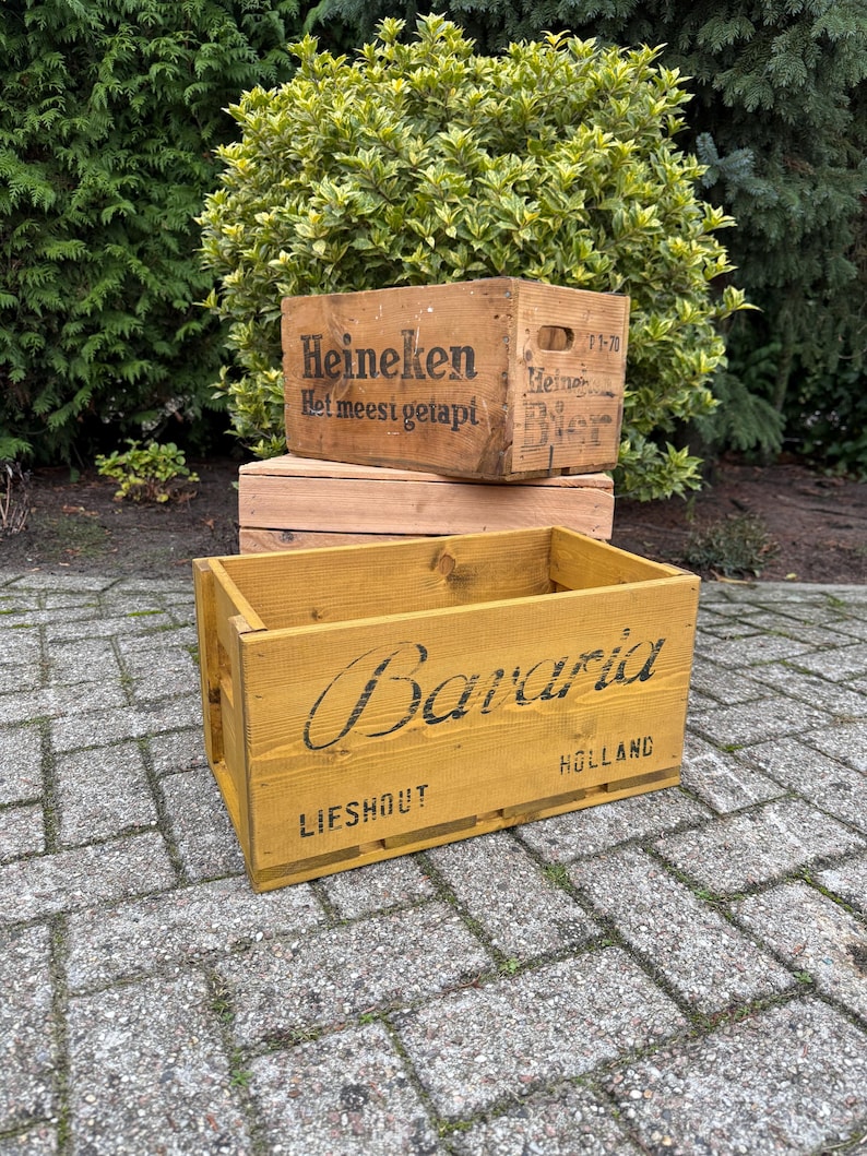 May include: Wooden crates with printed text. The top crate is light brown with the word "Heineken" and other text. The bottom crate is yellow with the word "Bavaria" and "Holland" printed on it. The crates are stacked on a brick surface.