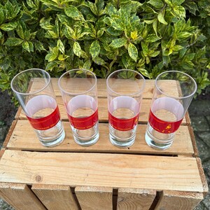 May include: Four clear drinking glasses with a red band and white text. The glasses are on a wooden crate, with green foliage behind. The glasses are cylindrical and empty. The text on the band is visible.