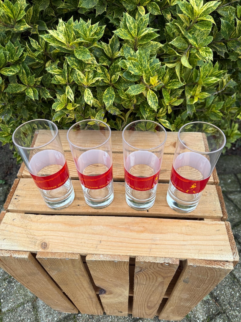 May include: Four clear drinking glasses with a red band and white text. The glasses are on a wooden crate, with green foliage behind. The glasses are cylindrical and empty. The text on the band is visible.