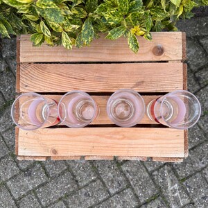 May include: Four clear drinking glasses with a pink stripe design, arranged on a wooden pallet. The glasses are empty and sit on a light brown wooden surface. Green and yellow foliage is visible in the background.