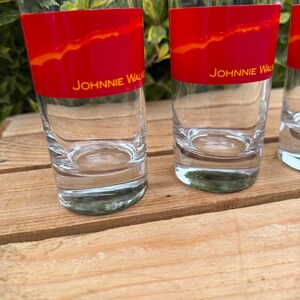 May include: Clear glass tumblers with a red band featuring an orange design and the text "JOHNNIE WALKER". The glasses are cylindrical and sit on a wooden surface. The background includes green foliage.