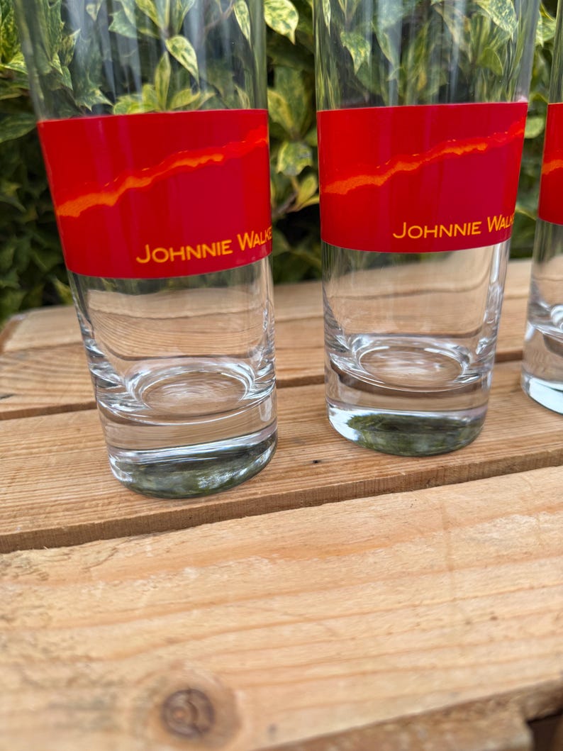 May include: Clear glass tumblers with a red band featuring an orange design and the text "JOHNNIE WALKER". The glasses are cylindrical and sit on a wooden surface. The background includes green foliage.