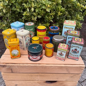 Può includere: Una collezione di barattoli di metallo vintage di varie forme e dimensioni, tra cui rotondi e rettangolari. I barattoli presentano disegni colorati e testo, come "Cacao", "Maggi" e "Droste's Cacao". Sono disposti su una cassa di legno.