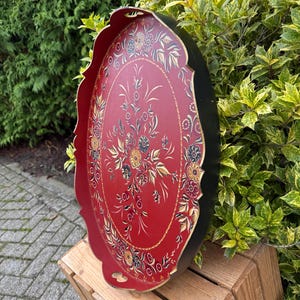 May include: Oval serving tray with a red interior and black exterior. The tray features a floral design in gold, white, and other colours. The tray has a scalloped edge and is resting on a wooden crate.