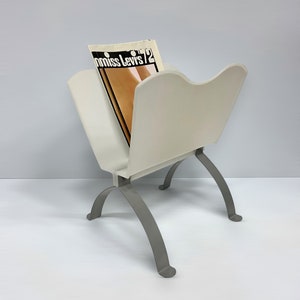 May include: White magazine rack with silver metal legs. The rack is holding a magazine with the text "miss Levis '72" on the cover.