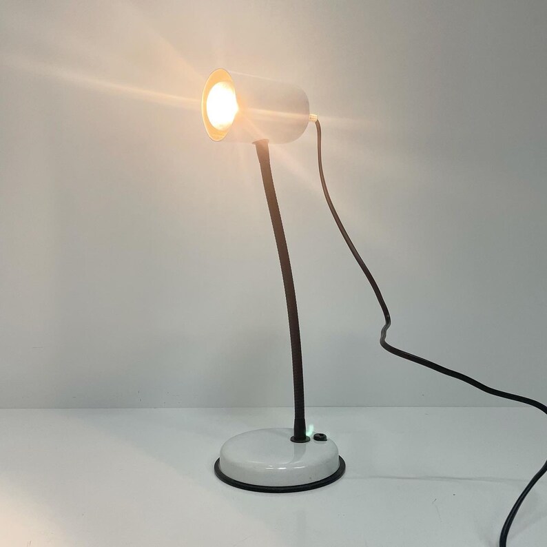 May include: A white desk lamp with a black cord and a white cylindrical shade. The lamp is turned on and the light is shining.