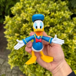Pode incluir: Uma figura do Pato Donald, com uma camisa de marinheiro azul, chap&eacute;u e p&eacute;s amarelos. A figura tem luvas brancas e uma gravata borboleta vermelha. A personagem de desenho animado posa com os bra&ccedil;os estendidos.
