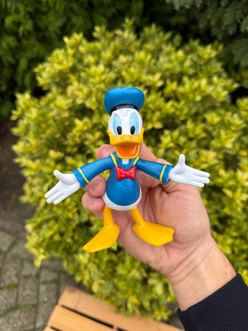 Pode incluir: Uma figura do Pato Donald, com uma camisa de marinheiro azul, chap&eacute;u e p&eacute;s amarelos. A figura tem luvas brancas e uma gravata borboleta vermelha. A personagem de desenho animado posa com os bra&ccedil;os estendidos.