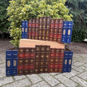 May include: A collection of decorative book sculptures. The books are in various colours, including blue, brown, and red. The spines feature gold lettering and decorative patterns. The books are arranged on a wooden surface, creating a unique display.