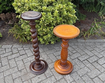 Puede incluir: Dos soportes de madera para plantas. Uno es de color marrón oscuro con un tallo en espiral, el otro es de color marrón claro con un tallo torneado. Ambos tienen tapas y bases redondas. Se exhiben al aire libre sobre un fondo de follaje verde.