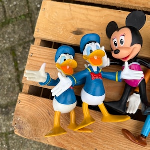 Pode incluir: Uma cole&ccedil;&atilde;o de figuras de personagens da Disney, incluindo duas figuras do Pato Donald em trajes e chap&eacute;us de marinheiro azuis, uma figura da Minnie Mouse com saia rosa e outros personagens. As figuras est&atilde;o em uma superf&iacute;cie de madeira.
