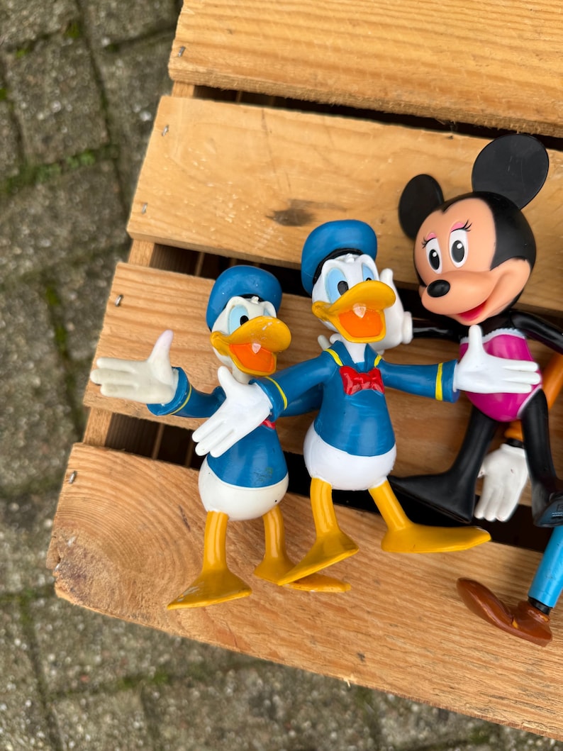 Pode incluir: Uma cole&ccedil;&atilde;o de figuras de personagens da Disney, incluindo duas figuras do Pato Donald em trajes e chap&eacute;us de marinheiro azuis, uma figura da Minnie Mouse com saia rosa e outros personagens. As figuras est&atilde;o em uma superf&iacute;cie de madeira.
