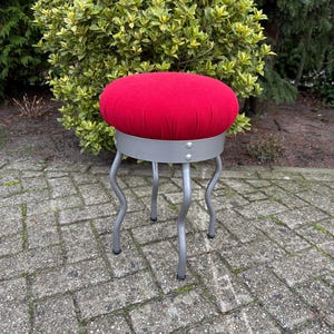 May include: A red upholstered stool with a round seat and a silver metal base. The stool has four curved legs and is set on a brick patio. The seat is approximately 14 inches in diameter.