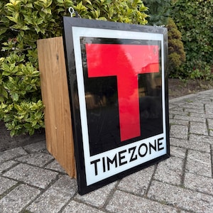 以下が含まれることがあります： 黒いフレームと白いブロック文字で「TIMEZONE」という文字が入った長方形の看板。看板は、黒い背景に大きな赤い「T」と白い縁取りが特徴です。上部には金属製のフックが付いています。