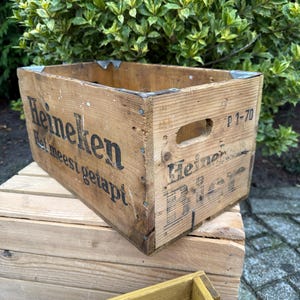 May include: A vintage wooden Heineken crate, showing signs of wear. The crate features the Heineken logo and text, metal corner reinforcements, and a handle. The wood has a natural, aged look.