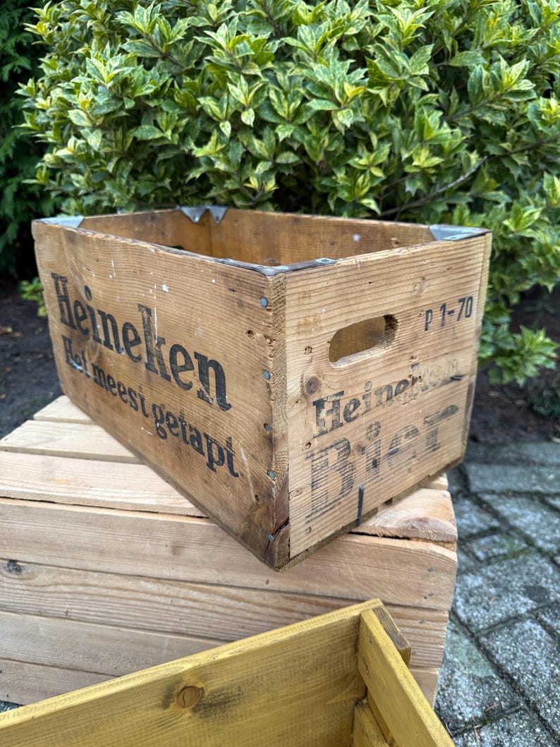 May include: A vintage wooden Heineken crate, showing signs of wear. The crate features the Heineken logo and text, metal corner reinforcements, and a handle. The wood has a natural, aged look.