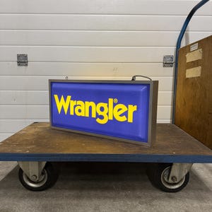 May include: A rectangular sign with the word "Wrangler" in yellow letters on a blue background. The sign is in a brown frame and sits on a blue and brown cart with wheels.