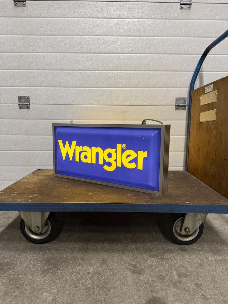 May include: A rectangular sign with the word "Wrangler" in yellow letters on a blue background. The sign is in a brown frame and sits on a blue and brown cart with wheels.