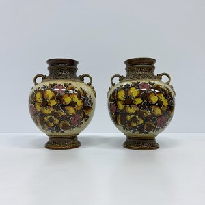 May include: Two brown ceramic vases with floral designs. The vases have a cream background with yellow, brown, and pink flowers. The vases have ornate brown trim and two handles.