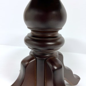 May include: A close-up of a dark brown wooden table base. The base has a turned, baluster-shaped central column and three curved legs. The wood has a glossy finish, and the background is white.