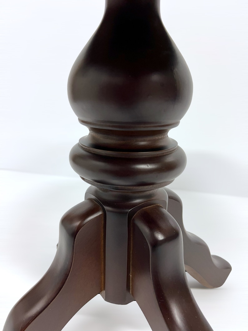 May include: A close-up of a dark brown wooden table base. The base has a turned, baluster-shaped central column and three curved legs. The wood has a glossy finish, and the background is white.