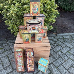 Puede incluir: Una colección de latas de metal vintage, apiladas sobre una caja de madera. Las latas presentan ilustraciones coloridas y texto, con una variedad de formas y tamaños. Las latas son en tonos de marrón, verde y azul, con ilustraciones detalladas.