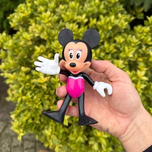 Pode incluir: Uma figura da Minnie Mouse com orelhas pretas, uma roupa rosa e branca e luvas brancas. A figura est&aacute; em pose com um bra&ccedil;o levantado, em um fundo de arbusto verde.