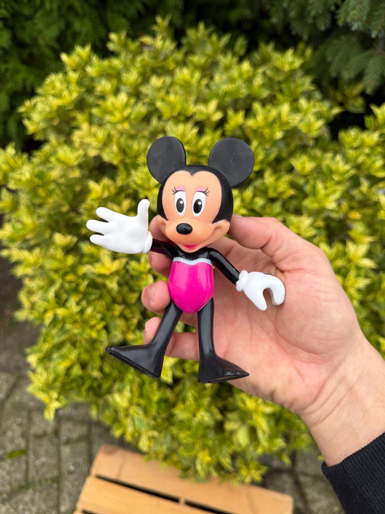 Pode incluir: Uma figura da Minnie Mouse com orelhas pretas, uma roupa rosa e branca e luvas brancas. A figura est&aacute; em pose com um bra&ccedil;o levantado, em um fundo de arbusto verde.