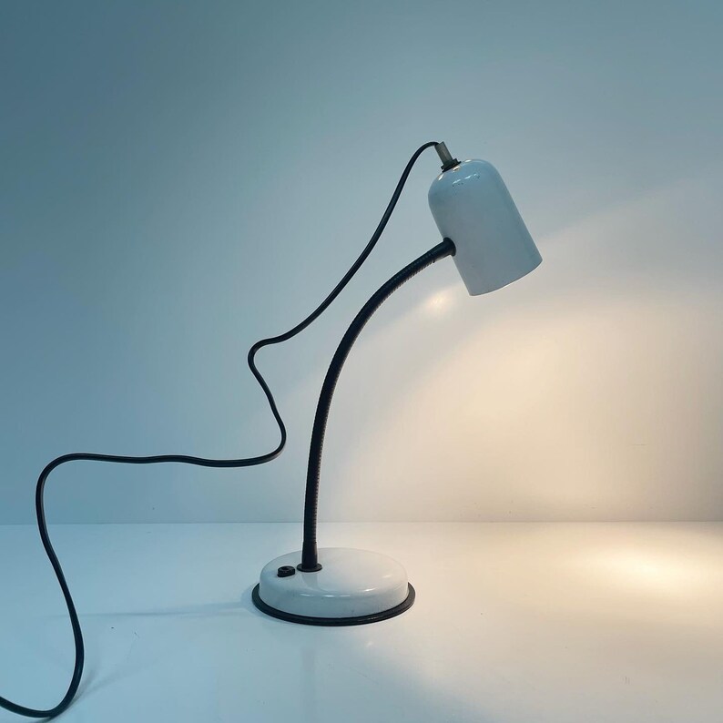 May include: A white desk lamp with a flexible black arm and a white shade. The lamp has a white base and a black cord.