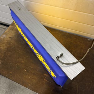 May include: A rectangular sign with the word "Wrangler" in yellow letters on a blue background. The sign has a silver metal frame and a power cord. The sign is resting on a brown surface.