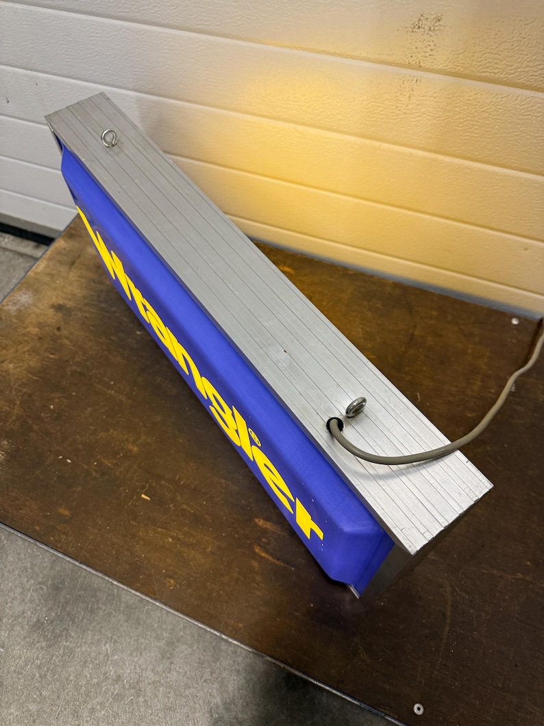 May include: A rectangular sign with the word "Wrangler" in yellow letters on a blue background. The sign has a silver metal frame and a power cord. The sign is resting on a brown surface.